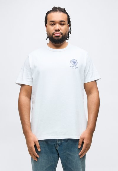 Άνδρας με πλεξούδες, που φοράει λευκό μπλουζάκι Ralph Lauren και μπλε τζιν, στέκεται όρθιος μπροστά σε ένα απλό ανοιχτόχρωμο φόντο.