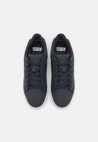 Sneakers azul escuro com material texturizado, apresentando uma sola de borracha branca, design com atacadores e detalhes em vermelho no interior e no logotipo lateral.