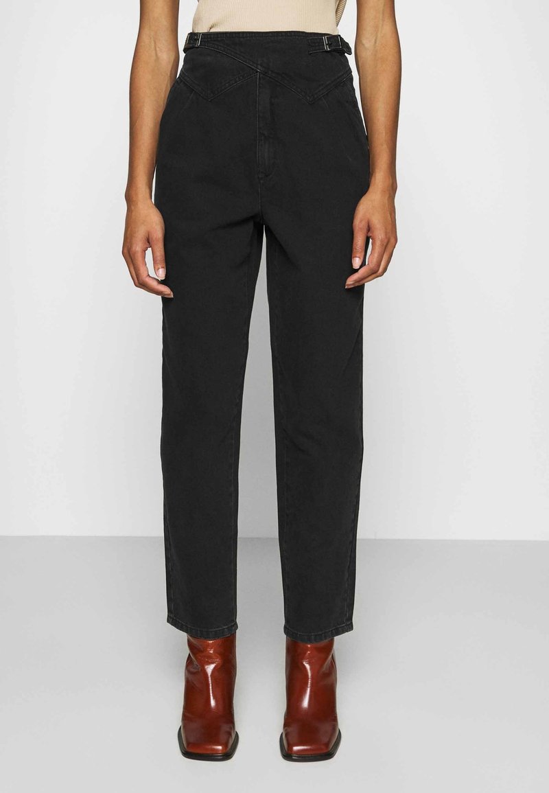 Pantalones de mezclilla negros de talle alto con un diseño de pierna recta, que presentan una costura frontal única y lazos para el cinturón, combinados con botas de tobillo rojas y puntiagudas.