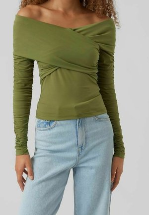 Frau trägt ein olivgrünes, schulterfreies, gerafftes Langarmoberteil in Kombination mit hellblauen, hoch taillierten Jeans.