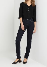 Kvinde iført en sort V-hals langærmet bluse, mørke skinny jeans og sorte lavhælede slingback sko, som står foran en hvid baggrund.