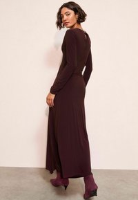 Rochie lungă, ajustată, de culoare burgundă, cu mâneci lungi, decolteu rotund și detaliu în formă de cheiță la spate, asortată cu cizme de gleznă în nuanță de prune. Textură de stofă netedă.