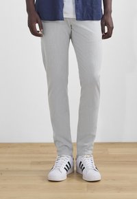 Lys grå, lige bukser lavet af struktureret stof, parret med hvide sneakers med sorte striber. Baggrunden er et trægulv.