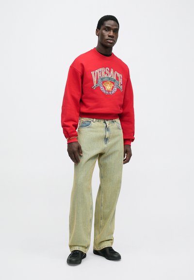 Punane sweatshirt, millel on "Versace" logo ja graafika, koos helesiniste triibuliste teksadega. Mustad jalanõud viimistlevad riietuse. Lihtne taust.
