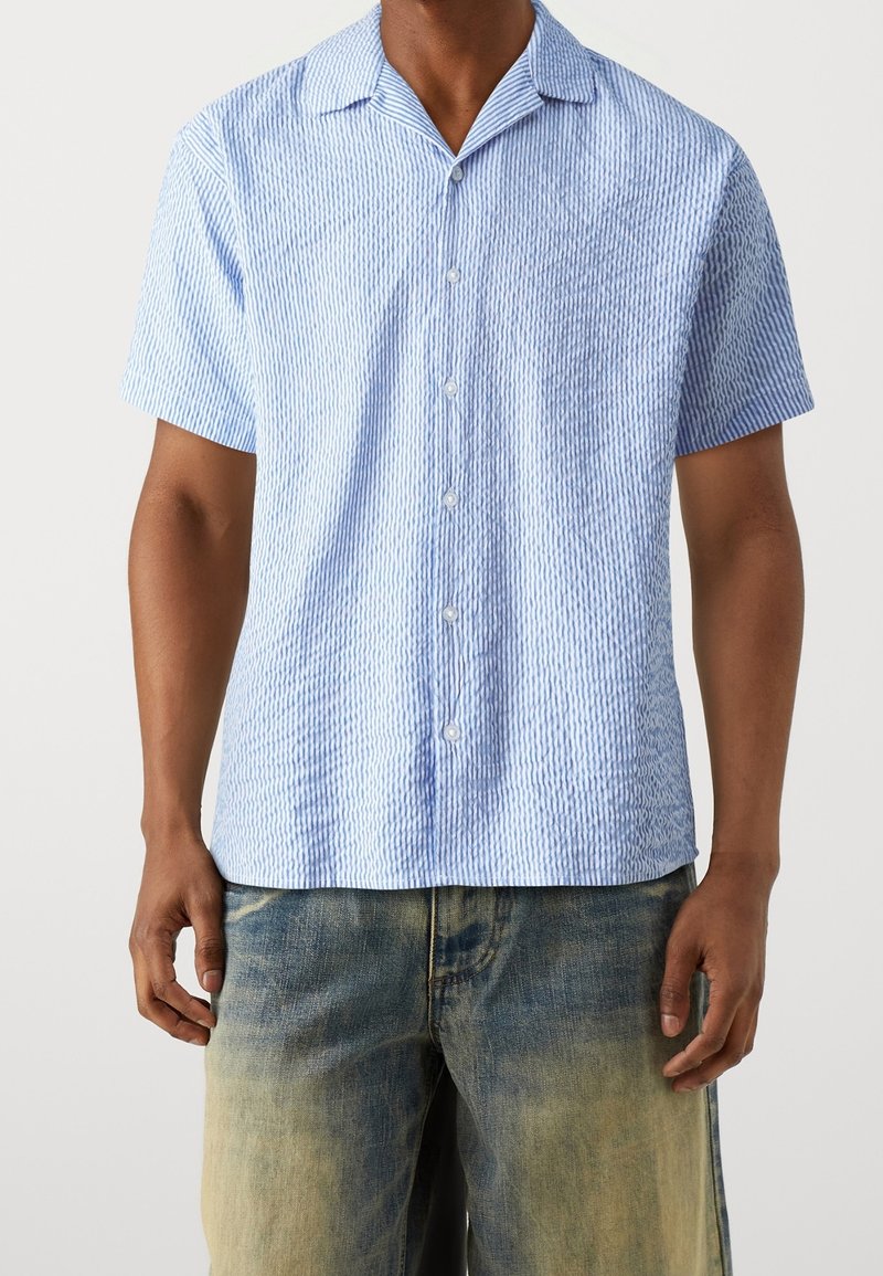 Homme portant une chemise à manches courtes bleu clair texturée avec col et un jean ample délavé.