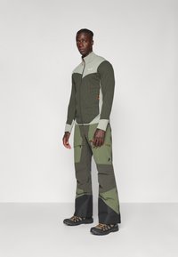Veste zippée verte et grise avec texture côtelée, assortie à un pantalon cargo multicolore. Comprend des poches zippées et des chaussures de randonnée noires robustes.