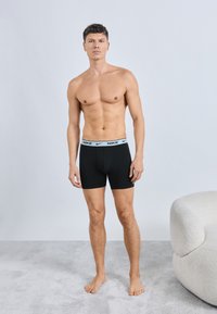 Fekete, Nike logóval díszített derékpántú boxeralsó. A modell ing nélkül, sima textúrával és testhezálló kialakítással áll a szőnyegen.