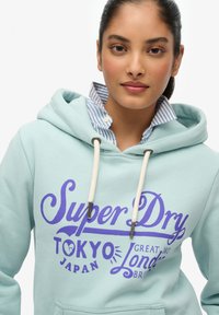 Turkusowy hoodie z fioletowym napisem "Super Dry", z kapturem na sznurku i kieszenią kangurką, nałożony na niebieską koszulę w paski.