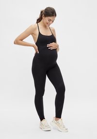 Body de maternidad negro con tirantes finos y un diseño ajustado, con una textura suave. Combinado con zapatillas blancas, frente a un fondo sencillo.