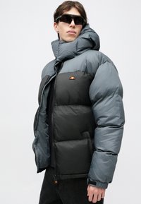 Pérová bunda v odtieňoch šedej s vysokým golierom, obsahujúca čiernu časť, predný zips a logo na ľavej strane hrudníka. Hladká, polstrovaná textúra.