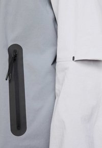 Prenda de color gris claro con un bolsillo vertical con cremallera de un gris más oscuro en la manga izquierda; textura suave, diseño minimalista.