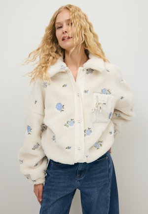 Vrouw met lang krullend blond haar, die een wit fleecejack met blauwe bloemmotieven en een blauwe spijkerbroek draagt tegen een effen achtergrond.
