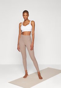 Soutien-gorge de sport blanc et leggings beiges à taille haute en matière côtelée. Le modèle se tient sur un tapis beige clair, pieds nus, avec les bras détendus.