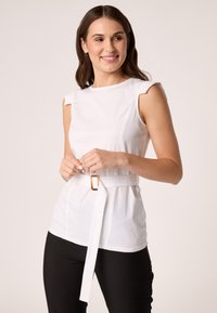 Weiße ärmellose Bluse mit taillierter Passform, breitem Gürtel mit goldener Schnalle und strukturiertem Stoff. Kombiniert mit schwarzen Slim-Fit-Hosen.