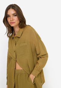 Χακί πουκάμισο σε υπερμεγέθη γραμμή με γιακά, κλείσιμο με κουμπιά και τσέπη στο στήθος· ελαφρύ ύφασμα με υφή και άνετη εφαρμογή.