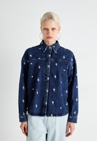 Camisa de ganga em azul escuro, com bordado floral branco, fecho em botão à frente, colarinho clássico e mangas longas com punhos em botão.