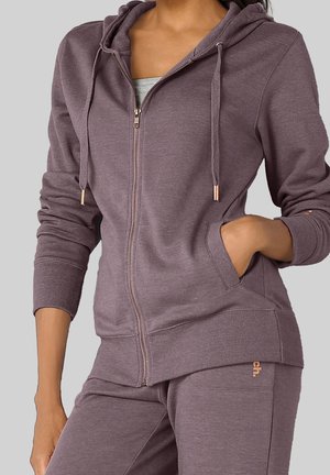Femme portant un sweat à capuche zippé mauve avec poches avant et un pantalon de survêtement assorti, main dans la poche, sur un fond gris uni.