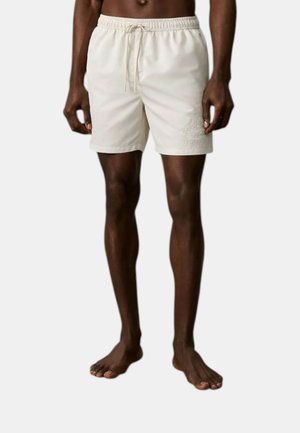 Persona dalla pelle scura che indossa pantaloncini beige con coulisse e logo ricamato sulla gamba sinistra, in piedi a piedi nudi su sfondo neutro.