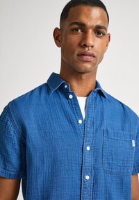 Camisa de manga corta, abotonada, en tejido azul texturizado. Cuenta con un bolsillo en el pecho y botones blancos, con un cuello plegado.