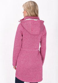 Fleece mēteli rozā tonī ar kapuci, garām piedurknēm un jostu viduklī. Iezīmēts ar izliekto apakšmalu un mīkstu tekstūru.