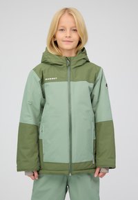 Bambino con capelli biondi alla spalle che indossa un giubbotto invernale Marmut verde bicolore e pantaloni abbinati, in piedi su uno sfondo semplice.