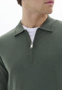 Pullover verde lavorato a maglia con colletto a zip, caratterizzato da una texture liscia. Indossato sopra una T-shirt bianca. Dettagli in metallo argentato sulla zip.