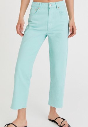 Lyseblå jeans med høj talje og lige ben med rå kant, båret med sorte flade sandaler med remme på en stående person.