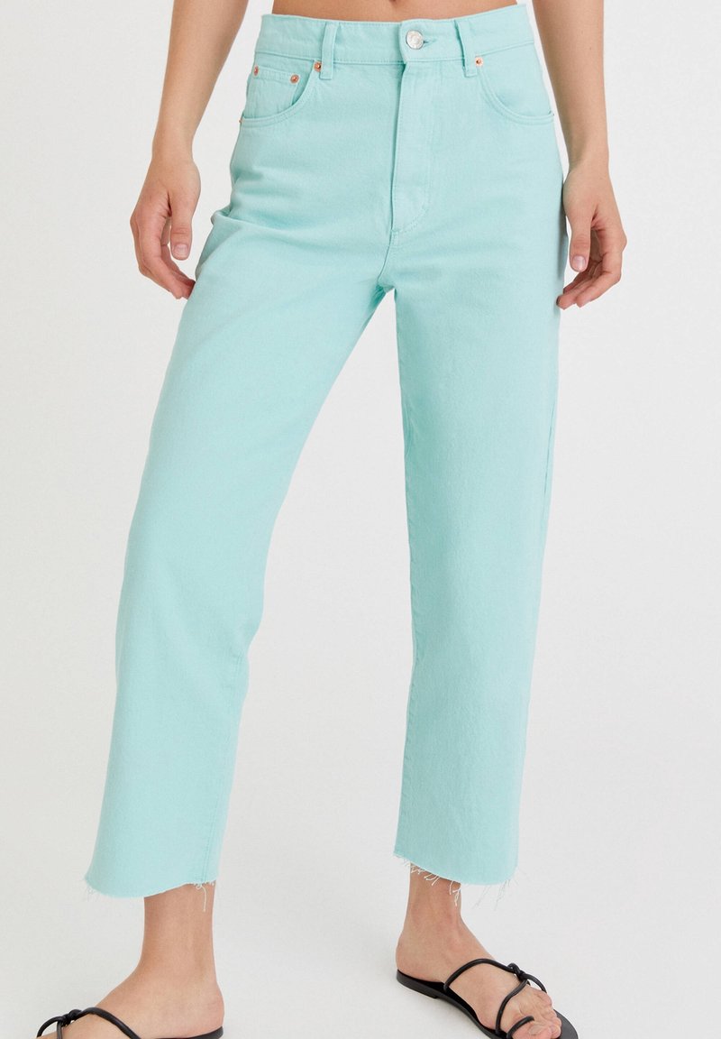 Jean bleu clair taille haute, coupe droite avec ourlet brut, porté avec des sandales plates noires à lanières sur une personne debout.