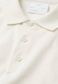 Polo beige en tissu texturé, avec un col à revers, trois boutons et une étiquette indiquant taille EU M, fabriqué au Bangladesh.