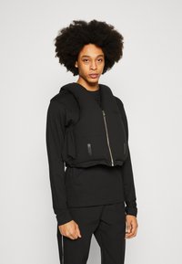 Colete preto cropped com fecho, apresenta frente acolchoada, dois bolsos frontais e tecido texturizado. Usado sobre uma camisola preta de manga comprida.