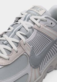 Κοντινό πλάνο του αθλητικού παπουτσιού Nike Vomero 5 σε ανοιχτό γκρι χρώμα, με λευκά κορδόνια και υφή στην πλαϊνή λεπτομέρεια.
