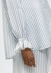 Κοντινό πλάνο ενός χεριού που φοράει λευκό πουκάμισο με μακριά μανίκια και παντελόνι με κάθετο μπλε ριγέ μοτίβο και ελαστικές λεπτομέρειες στα μανίκια.