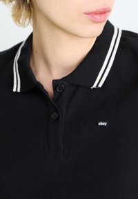 Čierny polokošeľ s textúrovanou tkaninou, s dvoma gombíkmi na légu, bielym pruhovaným golierom a malým logom "obey" na hrudi.