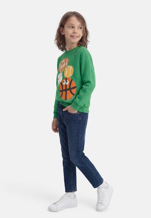 Bambino con felpa verde con palline sportive da cartone animato, jeans blu scuro, scarpe da ginnastica bianche, che guarda di lato con una mano in tasca.