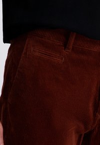 Brown corduroy pants with a fine ribbed texture, featuring a single back pocket and straight-cut design. Black shirt partially visible above.