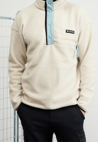 Pullover in pile color crema con colletto alto, zip nera e dettagli azzurri. Presenta una tasca sul petto con zip e un tessuto texturizzato.