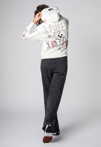 White hoodie featuring a teddy bear graphic and floral designs on the back, paired with black jeans and low-top sneakers with star accents.