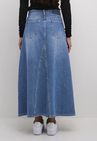 Falda maxi de mezclilla con un lavado azul, que presenta una costura trasera, dos bolsillos traseros y un corte recto. Combinada con zapatillas blancas.