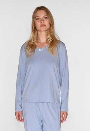Donna bionda che indossa un top per pigiama a maniche lunghe azzurro chiaro con rifiniture in pizzo e pantaloni da pigiama coordinati, in piedi su sfondo bianco.