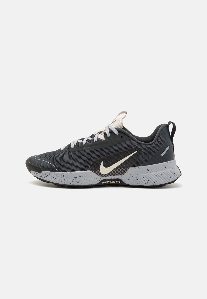 Chaussure de trail Nike noire et grise avec lacets blancs, semelle tachetée et logo Swoosh beige sur fond uni.