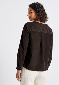 Vrouw met krullend haar, gekleed in een donkerbruine lange mouwtop en lichtkleurige broek, kijkt over haar schouder tegen een effen achtergrond.