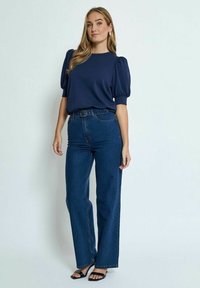 Marinblå puffärmad topp i kombination med högmidjade, mörkblå vida jeans; modell som bär svarta remsandaler och minimala accessoarer.