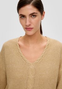 Beige gestrickter Pullover mit V-Ausschnitt, der ein strukturiertes Zopfmuster entlang des Ausschnitts sowie einen lockeren, entspannten Schnitt aufweist.