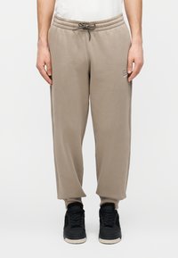 Beige sweatpants met een elastische tailleband en trekkoord, voorzien van zijzakken en een klein logo op de linker dij, gecombineerd met zwarte sneakers.