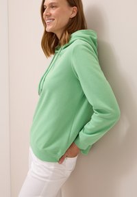 Felpa verde con cappuccio in morbido tessuto, dotata di tasca a marsupio, polsini a coste e vestibilità comoda. Indossata sopra pantaloni bianchi.