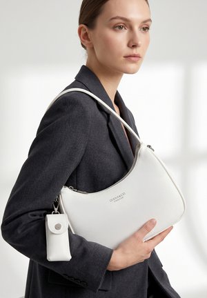 Femme en blazer noir tenant un sac à main blanc DIANA&CO Firenze avec une petite pochette assortie attachée, sur un fond neutre et lumineux.