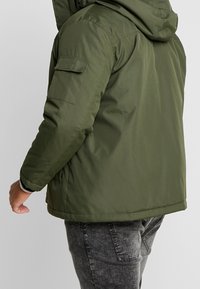 Mann in olivgrünem Kapuzenjacke mit Klappentasche am Ärmel und schwarzen gewaschenen Jeans, steht vor einem schlichten Hintergrund.