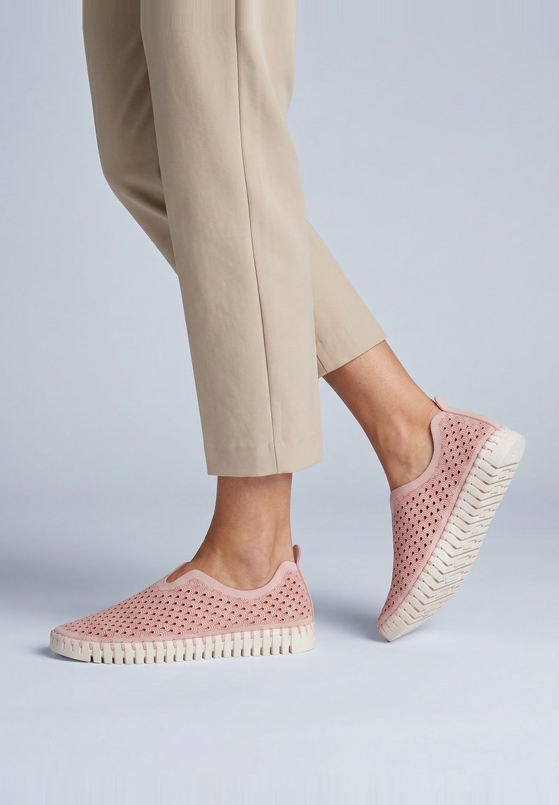 Persona que lleva pantalones beige recortados y zapatos rosas sin cordones con patrones perforados y suelas blancas gruesas dando un paso adelante.