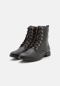 Un par de botas de cuero negro hasta el tobillo con ojales dorados y cordones negros sobre un fondo blanco.