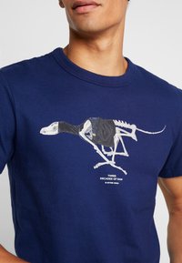 Námořnická modrá bavlněná tričko s grafikou kosterního ptáka v černé vestě, s bílým textem "TŘI DEKÁDY SYROVÉHO."